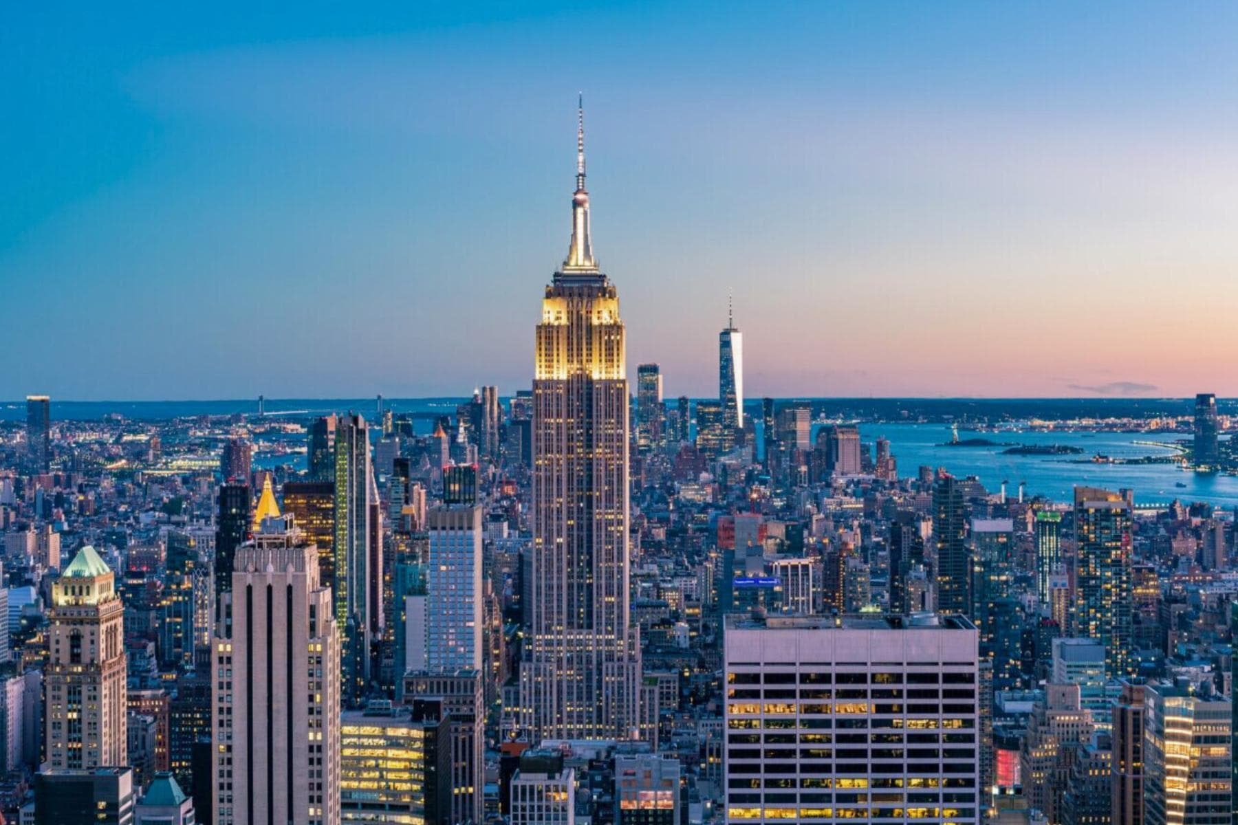 A wide city view at sunset, with many skyscrapers and the Empire State Building prominently featured.