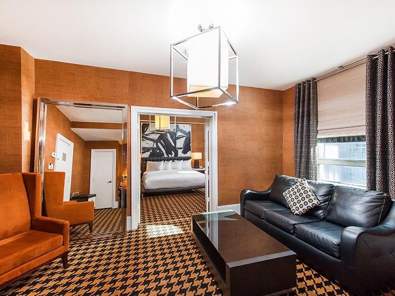 A wide view of a hotel room's living area with a black leather sofa, a glass coffee table, two armchairs, and a chandelier hanging from the ceiling.