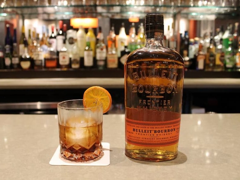 a glass of brown liquor on the rocks next to a bottle of Bulleit Bourbon