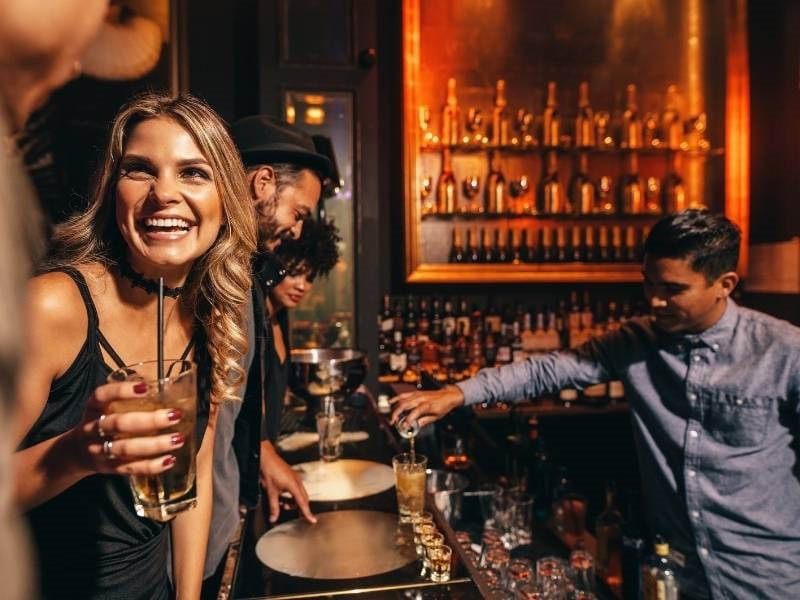 A woman smiles while holding a drink at a bar with a bartender mixing drinks.