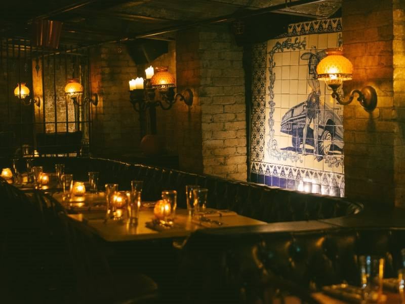 A dimly lit dining area with a long table set for multiple people, candles, and a tiled wall featuring a car image.