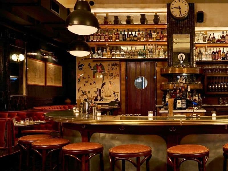 A dimly lit bar with a long counter, stools, and shelves of bottles in the background.
