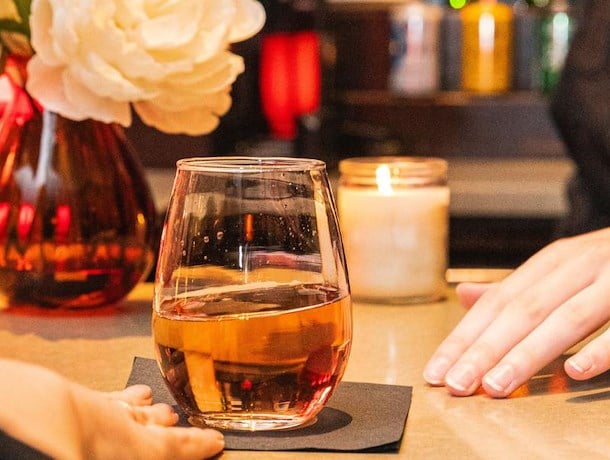 A glass of amber liquid on a bar with a lit candle and flowers in the background