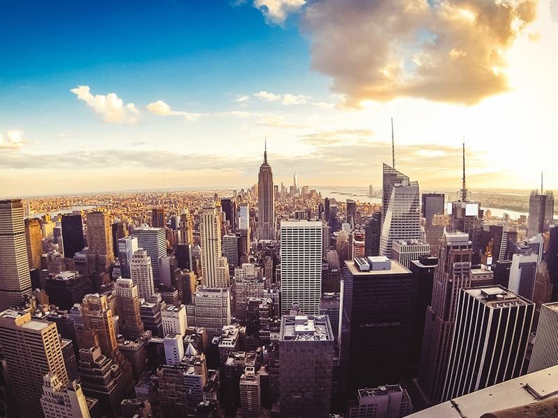 A wide-angle view of a cityscape with many skyscrapers and a bright sun in the background.